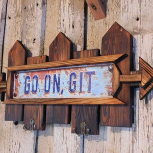 May include: A wooden sign with a rustic finish, shaped like a fence with an arrow pointing to the right. The sign has a metal plaque with the text "GO ON, GIT" in blue lettering on a white background. The sign has two hooks on the bottom.