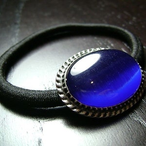 Op de afbeelding: Een zwarte haarband met een grote, ovale, donkerblauwe edelsteen in het midden. De edelsteen is gezet in een zilverkleurig, getextureerd metalen frame. De haarband ligt op een donker houten oppervlak.