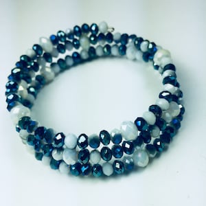 Op de afbeelding: Een wikkelarmband met rijen facetkralen in wit, lichtgrijs en diepblauw. De kralen zijn geregen op een flexibele draad, waardoor een spiraalvormig ontwerp ontstaat. De armband wikkelt zich meerdere keren om de pols.