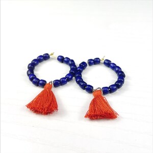 Op de afbeelding: Een paar oorringen. Elke oorring heeft een ring van donkerblauwe kralen met een feloranje kwast. De oorbellen worden gepresenteerd op een wit oppervlak.