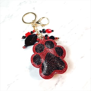 Puede incluir: Un llavero con forma de huella de pata roja y negra con pedrería. El llavero tiene una cadena dorada y un anillo dorado.