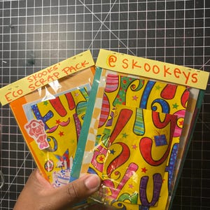 May include: Two colorful scrap packs are held in a hand. The packs have yellow tops with text that reads "Eco Scrap Pack" and "@skookeys". The packs contain patterned paper with red, blue, and green designs.