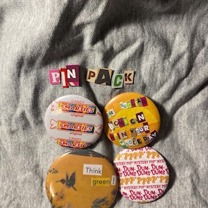 May include: A set of four circular pin-back buttons with various designs. One features Smarties candy branding, another reads One Nuclear Bomb Can Ruin Your Whole Day, one shows a hummingbird with the text Think green!, and one displays Dum-Dums Mystery Pop branding. The words PIN PACK are arranged above the buttons in a ransom-note style font.