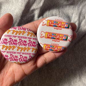 May include: Two round pinback buttons. One button features a white background with repeating red text reading DUM DUMS MYSTERY POP and yellow question marks. The other button is white with three red-outlined illustrations of Smarties candy rolls.