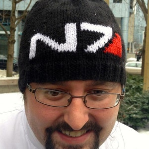 May include: A black knit beanie with a white knitted "N7" logo and a red heart on the side. The beanie is worn by a person with a beard and glasses.