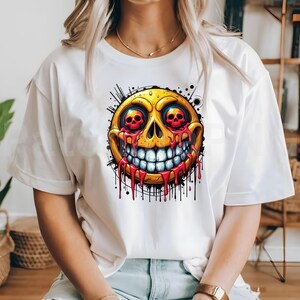 May include: White t-shirt featuring a vibrant, cartoonish design of a yellow emoji with red skull eyes and dripping red blood. The emoji has a wide, toothy grin and is surrounded by black splatter details.