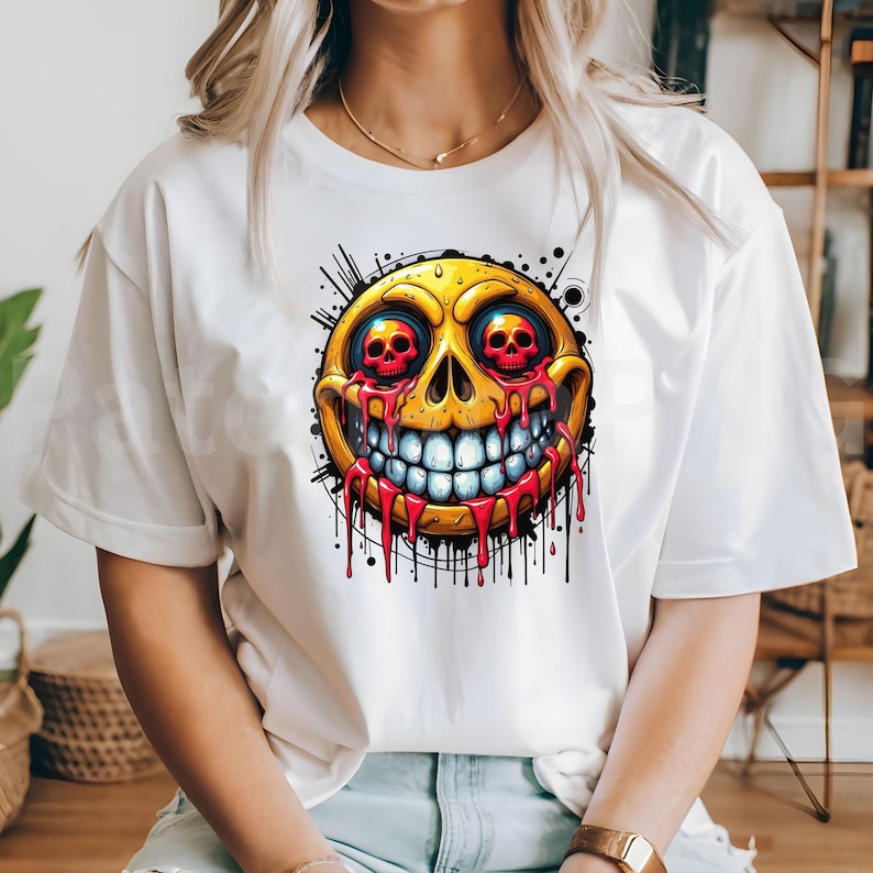 May include: White t-shirt featuring a vibrant, cartoonish design of a yellow emoji with red skull eyes and dripping red blood. The emoji has a wide, toothy grin and is surrounded by black splatter details.