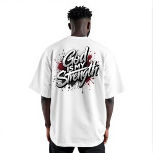 May include: White t-shirt with the text "God is my Strength" in a stylized font, accented by red and black paint splatter design. The shirt has short sleeves and a relaxed fit.
