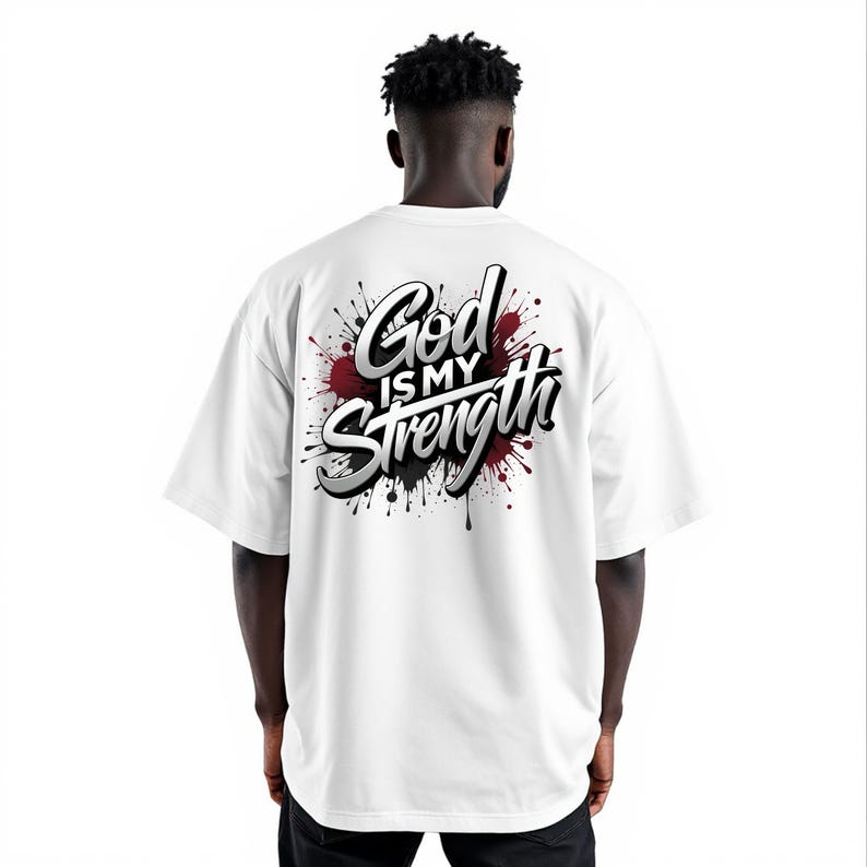 May include: White t-shirt with the text "God is my Strength" in a stylized font, accented by red and black paint splatter design. The shirt has short sleeves and a relaxed fit.