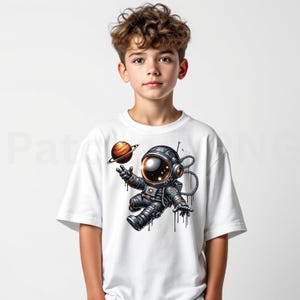 May include: White t-shirt featuring a cartoon astronaut floating in space, holding a planet. The astronaut is wearing a dark gray spacesuit with a reflective helmet. The planet is orange and brown with a ring around it. The design has a playful, illustrative style.