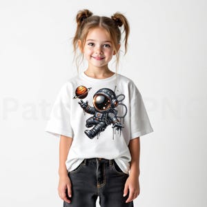 May include: White t-shirt featuring a cartoon astronaut in a spacesuit, holding a planet with rings. The astronaut is in a floating pose. The design is in shades of gray, black, orange, and brown.