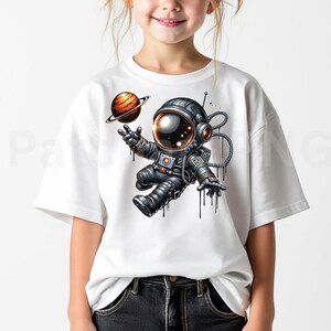 May include: White t-shirt featuring a cartoon astronaut in a spacesuit, with a planet in the background. The astronaut is in a floating pose, with black drips coming from the suit. The astronaut is wearing a helmet with a bright orange visor.