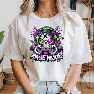 May include: White t-shirt featuring a skull wearing headphones and holding a game controller. The skull has a purple and green design, with the text "RAGE MODE" below. The design includes purple paint splatters.
