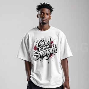 May include: White t-shirt with the text "God is my Strength" in a stylized, 3D design. The text is set against a backdrop of black and red paint splatters, creating a bold and eye-catching graphic.
