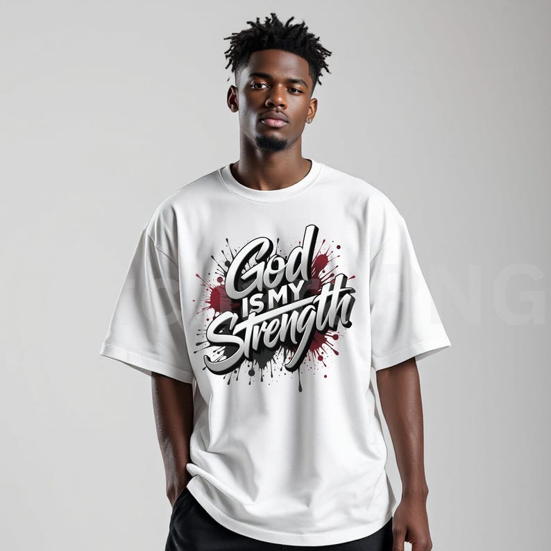 May include: White t-shirt with the text "God is my Strength" in a stylized, 3D design. The text is set against a backdrop of black and red paint splatters, creating a bold and eye-catching graphic.