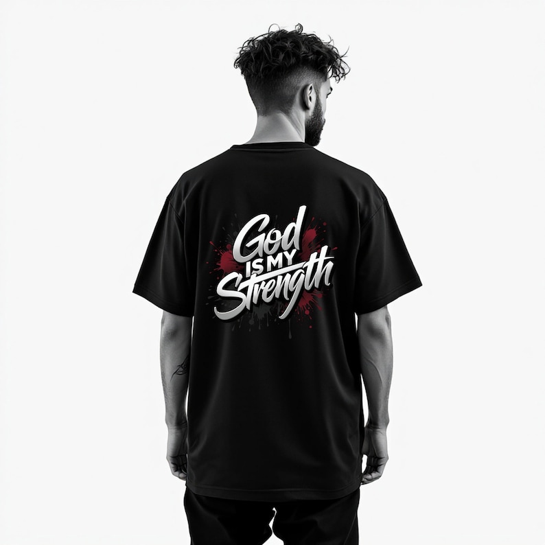 May include: Black t-shirt with the text "God is my Strength" in white and silver lettering, set against a red splatter design. The shirt is a crew neck style.