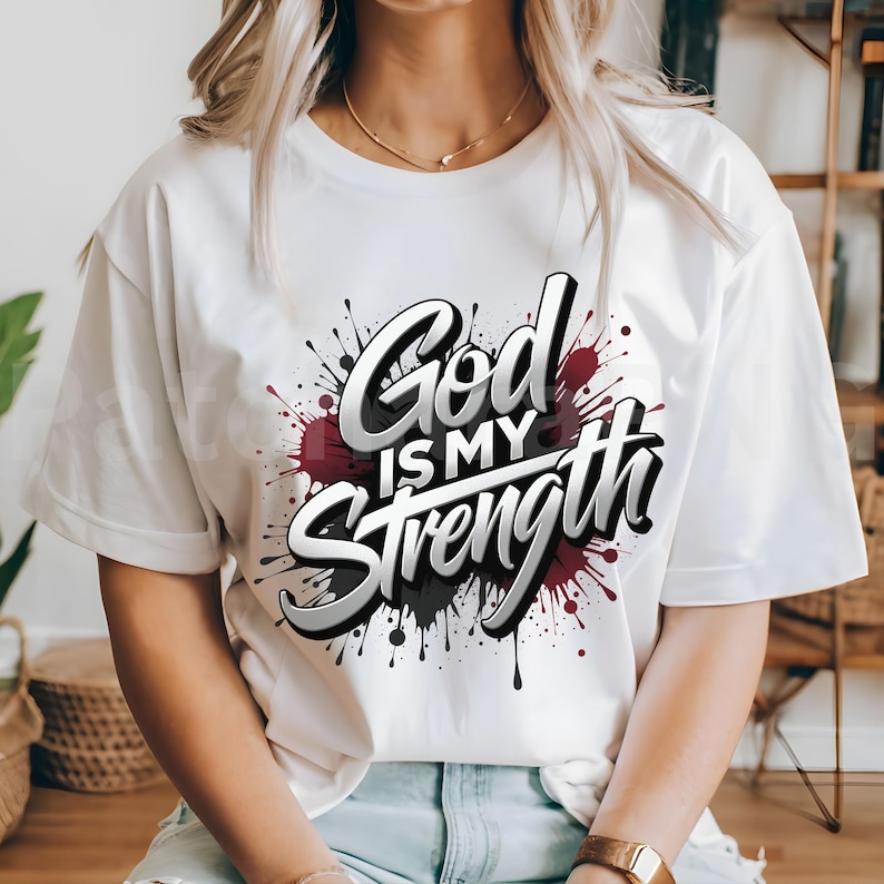 May include: White t-shirt with the text "God is my Strength" in a stylized, 3D font. The text is set against a backdrop of maroon, black, and gray paint splatters, creating a bold and eye-catching design.