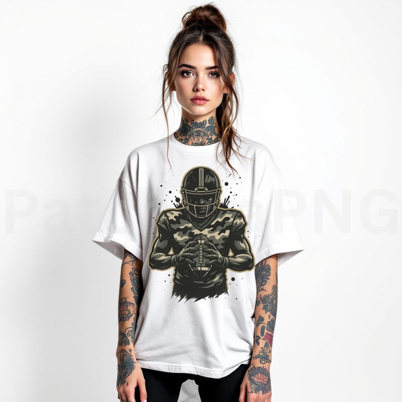 May include: White t-shirt featuring a football player in a helmet and camouflage uniform, holding a football. The graphic is in shades of green, brown, and black. The design has a sporty theme.