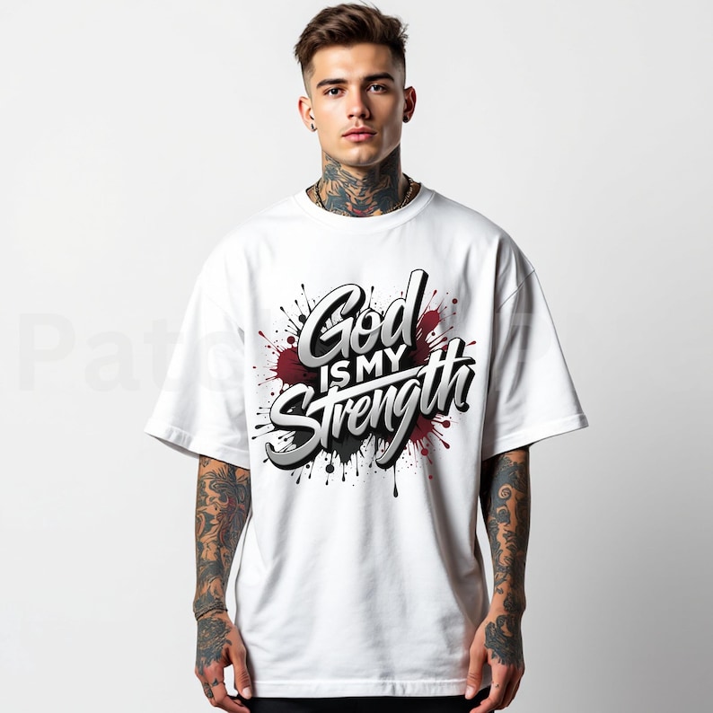 May include: White t-shirt with the text "God is my Strength" in a stylized font, set against a backdrop of red and black paint splatters. The shirt has short sleeves and a relaxed fit.