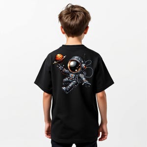 May include: Black t-shirt featuring a cartoon astronaut floating in space, with a planet in the background. The astronaut is wearing a detailed spacesuit with a reflective visor. The design is on the back of the shirt.