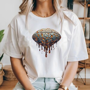 K&ouml;nnte beinhalten: Wei&szlig;es T-Shirt mit einem stilisierten Football-Design. Der Football hat eine rissige, lavaartige Textur in Blau-, Orange- und Braunt&ouml;nen, mit tropfendem geschmolzenem Material. Ein einzigartiges Grafik-T-Shirt.