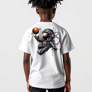 May include: White t-shirt featuring a colorful illustration of an astronaut floating in space with a planet. The astronaut is wearing a black and silver spacesuit. The design includes a planet with rings.