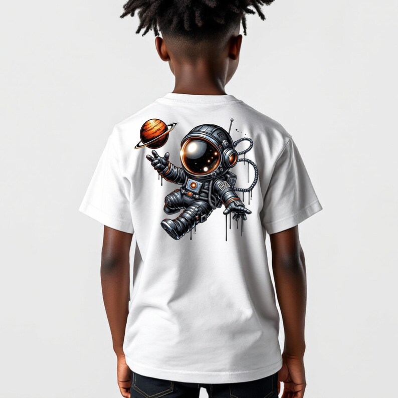 May include: White t-shirt featuring a colorful illustration of an astronaut floating in space with a planet. The astronaut is wearing a black and silver spacesuit. The design includes a planet with rings.