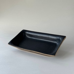 May include: A rectangular black ceramic tray with a slightly curved shape. The interior is a deep black, contrasting with the lighter, unglazed rim. The tray's design is simple and modern, suitable for serving or display.