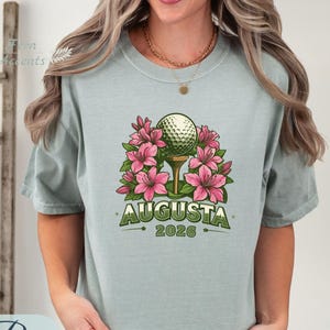 Pu&ograve; includere: T-shirt verde chiaro con una pallina da golf su un tee, circondata da fiori rosa. Il testo "AUGUSTA 2026" &egrave; stampato sotto. La maglietta &egrave; realizzata in un materiale morbido.