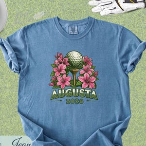 Pu&ograve; includere: T-shirt blu jeans con grafica a tema golf. Il design presenta una pallina da golf su un tee circondata da fiori rosa, con la scritta "Augusta" e l'anno "2026" in verde. Accessori: visiera bianca, palline da golf, tee e guanto da golf.
