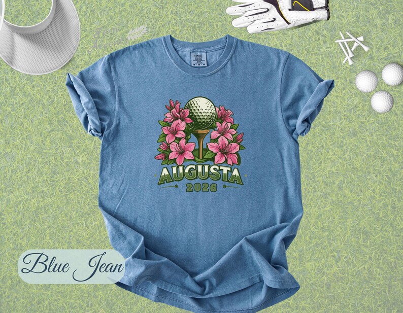 Pu&ograve; includere: T-shirt blu jeans con grafica a tema golf. Il design presenta una pallina da golf su un tee circondata da fiori rosa, con la scritta "Augusta" e l'anno "2026" in verde. Accessori: visiera bianca, palline da golf, tee e guanto da golf.