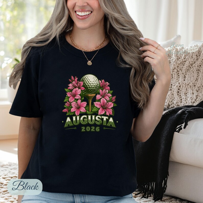 Pu&ograve; includere: T-shirt nero con una pallina da golf su un tee circondata da fiori rosa. Il testo "AUGUSTA 2026" &egrave; stampato sotto. La maglietta ha un girocollo.