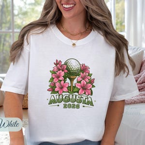 Pu&ograve; includere: T-shirt bianca con una pallina da golf su un tee, circondata da fiori rosa. Il design include la parola "AUGUSTA" e l'anno "2026" in verde. &Egrave; visibile una collana a catena dorata.
