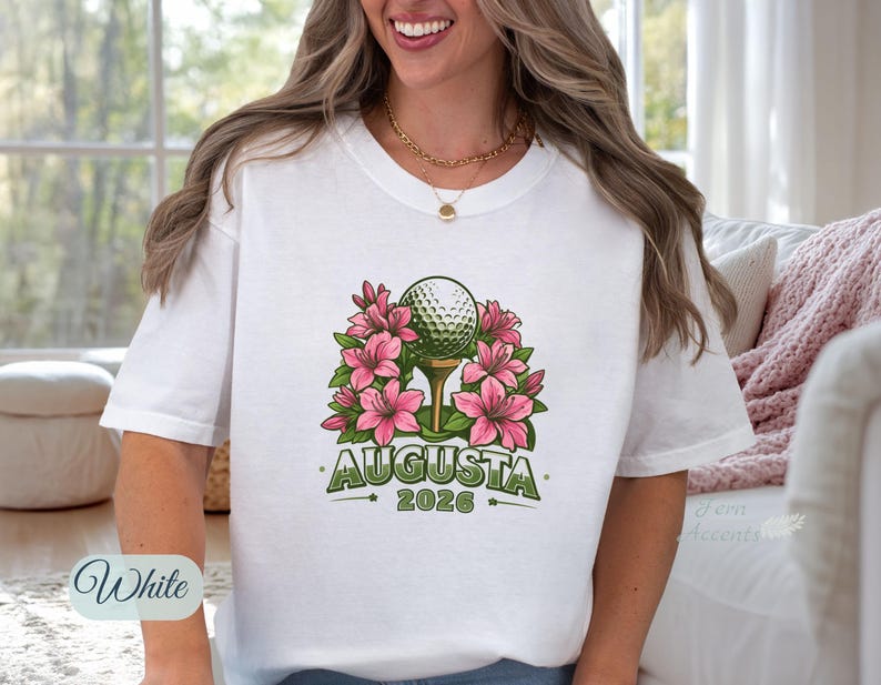 Pu&ograve; includere: T-shirt bianca con una pallina da golf su un tee, circondata da fiori rosa. Il design include la parola "AUGUSTA" e l'anno "2026" in verde. &Egrave; visibile una collana a catena dorata.