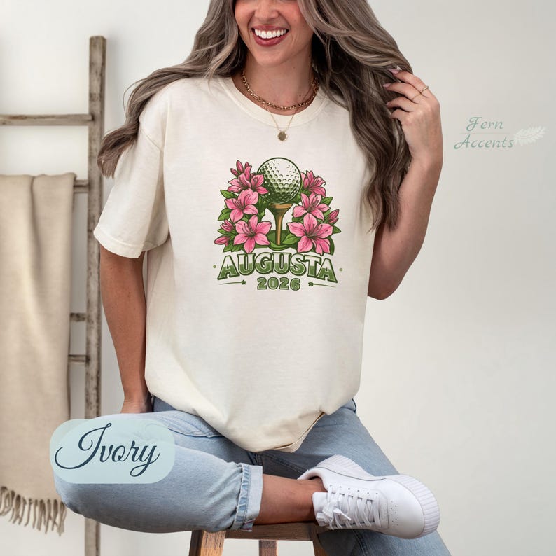 Pu&ograve; includere: T-shirt color avorio con una pallina da golf su un tee circondata da fiori rosa. La parola "AUGUSTA" &egrave; stampata in verde, con "2026" sotto. La maglietta &egrave; in stile casual.