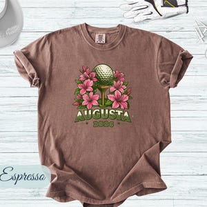 Pu&ograve; includere: T-shirt color espresso con una pallina da golf su un tee, circondata da fiori rosa. La parola "AUGUSTA" &egrave; arcuata sopra la pallina, con "2026" sotto. Accessori da golf sullo sfondo.