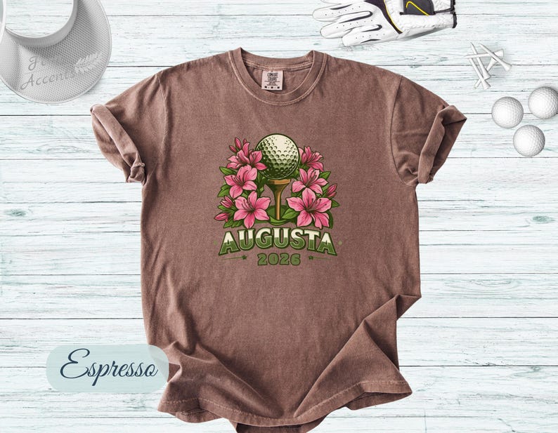 Pu&ograve; includere: T-shirt color espresso con una pallina da golf su un tee, circondata da fiori rosa. La parola "AUGUSTA" &egrave; arcuata sopra la pallina, con "2026" sotto. Accessori da golf sullo sfondo.