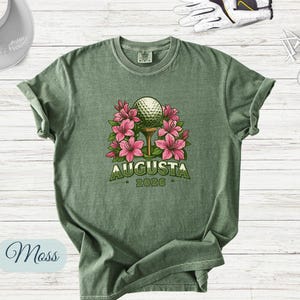 Pu&ograve; includere: T-shirt verde muschio con una pallina da golf su un tee, circondata da fiori rosa. La parola "Augusta" &egrave; stampata sopra l'anno "2026" in verde. Sono visibili una visiera da golf, palline da golf, tee e un guanto da golf.