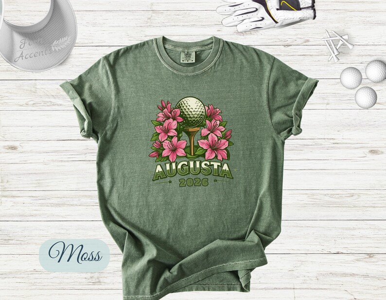 Pu&ograve; includere: T-shirt verde muschio con una pallina da golf su un tee, circondata da fiori rosa. La parola "Augusta" &egrave; stampata sopra l'anno "2026" in verde. Sono visibili una visiera da golf, palline da golf, tee e un guanto da golf.