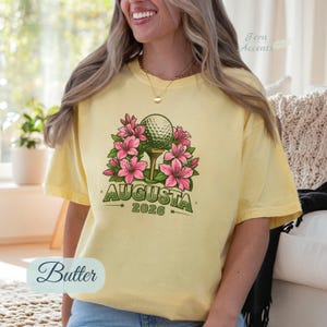Pu&ograve; includere: T-shirt giallo chiaro con una pallina da golf su un tee, circondata da fiori rosa. Il testo "AUGUSTA 2026" &egrave; stampato sotto. La maglietta &egrave; di colore giallo chiaro.