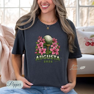 Pu&ograve; includere: T-shirt grigio scuro con una pallina da golf su un tee, circondata da fiori rosa. La parola "AUGUSTA" &egrave; stampata in verde, con "2026" sotto. La maglietta &egrave; in stile casual.