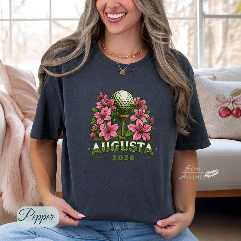 Pu&ograve; includere: T-shirt grigio scuro con una pallina da golf su un tee, circondata da fiori rosa. La parola "AUGUSTA" &egrave; stampata in verde, con "2026" sotto. La maglietta &egrave; in stile casual.