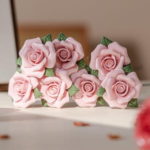 Pode incluir: Um par de brincos de cerâmica rosa em forma de rosas. As rosas estão dispostas em cacho e têm folhas verdes.