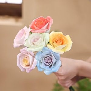 May include: A bouquet of colorful roses in shades of pink, orange, yellow, green, and blue. The roses are held in a hand, showcasing their vibrant colors and delicate petals. The flowers are a mix of pastel and bright hues.