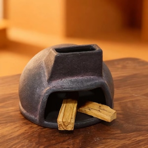 Puede incluir: Un porta incienso de cerámica gris oscuro con forma redondeada y una abertura rectangular en la parte superior. Dos palitos de palo santo de color marrón claro se colocan dentro del soporte. El soporte está sobre una superficie de madera.