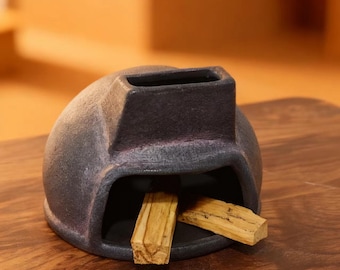 Pode incluir: Um porta-incenso de cerâmica cinza escuro com formato arredondado e uma abertura retangular na parte superior. Dois palitos de palo santo marrons claros são colocados dentro do suporte. O suporte está sobre uma superfície de madeira.