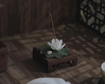 May include: A wooden incense holder featuring a white lotus flower and green leaf. An incense stick is lit, with smoke rising. The holder is placed on a wooden surface, creating a serene atmosphere, ideal for meditation.