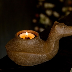 May include: A brown ceramic camel-shaped candle holder with a lit candle inside. The candle holder has a textured surface and a small opening for the candle. The camel's head is tilted slightly upward, and its eyes are closed.