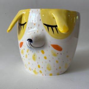 Op de afbeelding: Een keramische mok in de vorm van een hondenkop met gele oren, gesloten ogen met lange zwarte wimpers en een verhoogde neus. Het witte oppervlak is voorzien van gele en oranje vlekken, met een klein blauw detail op de neus van dit drinkgerei.