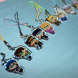 Puede incluir: Una colección de colgantes de personajes de estilo anime con diseños coloridos. Cada colgante presenta un personaje diferente con colores vibrantes y obras de arte detalladas. Los colgantes están unidos a llaveros plateados con cordones coloridos.